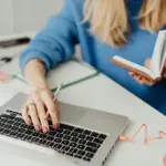 Frau mit blauen Pulli und Buch in der hand vor Laptop